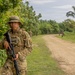 A uniformed soldier wearing multicam camouflage and a boonie hat stands at the edge of a dirt road in a tropical environment, holding an assault rifle. A name tape reading 'NAVAL' is visible on the chest, along with a unit patch on the sleeve. Additional soldiers are visible in the background along the road.