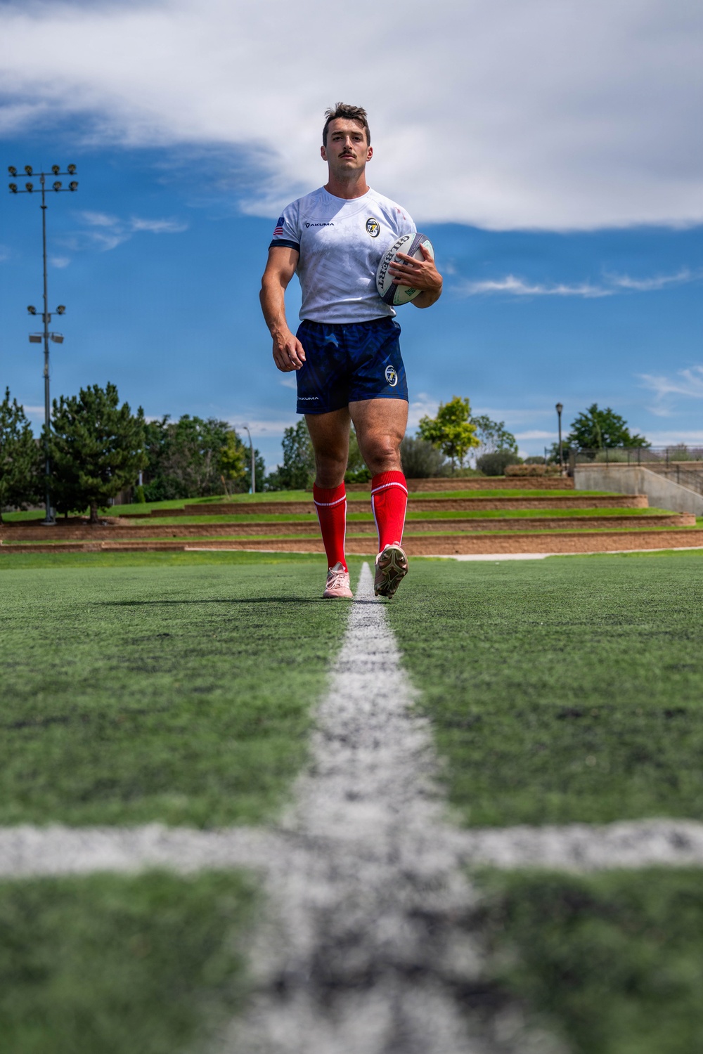 A male rugby player walks along a white line on an artificial turf field, holding a rugby ball in his right hand. He is wearing a white jersey with a US flag patch and team logo, navy blue shorts, red knee-high socks, and cleats. The photo is taken from a low ground-level angle, giving a dramatic perspective against a partly cloudy blue sky.