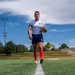 A male rugby player walks along a white line on an artificial turf field, holding a rugby ball in his right hand. He is wearing a white jersey with a US flag patch and team logo, navy blue shorts, red knee-high socks, and cleats. The photo is taken from a low ground-level angle, giving a dramatic perspective against a partly cloudy blue sky.