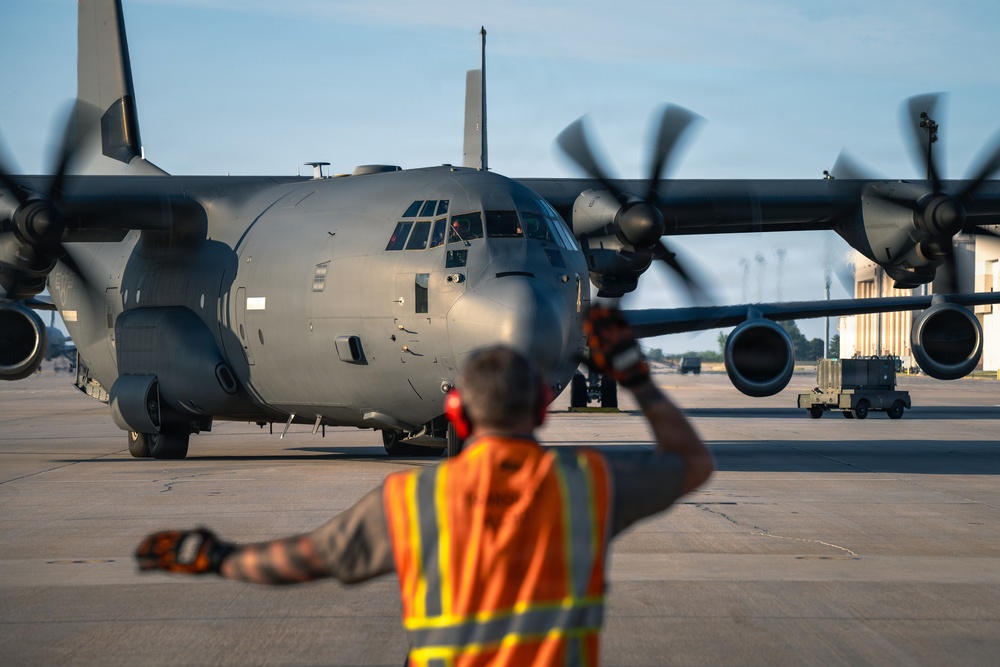 A ground crew member wearing an orange high-visibility safety vest is marshaling a large military turboprop aircraft on a flight line apron. The aircraft appears to be a Lockheed C-130 Hercules in gray military livery with all four propellers spinning. The image is taken during golden hour lighting, likely early morning or late evening.