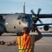 A ground crew member wearing an orange high-visibility safety vest is marshaling a large military turboprop aircraft on a flight line apron. The aircraft appears to be a Lockheed C-130 Hercules in gray military livery with all four propellers spinning. The image is taken during golden hour lighting, likely early morning or late evening.