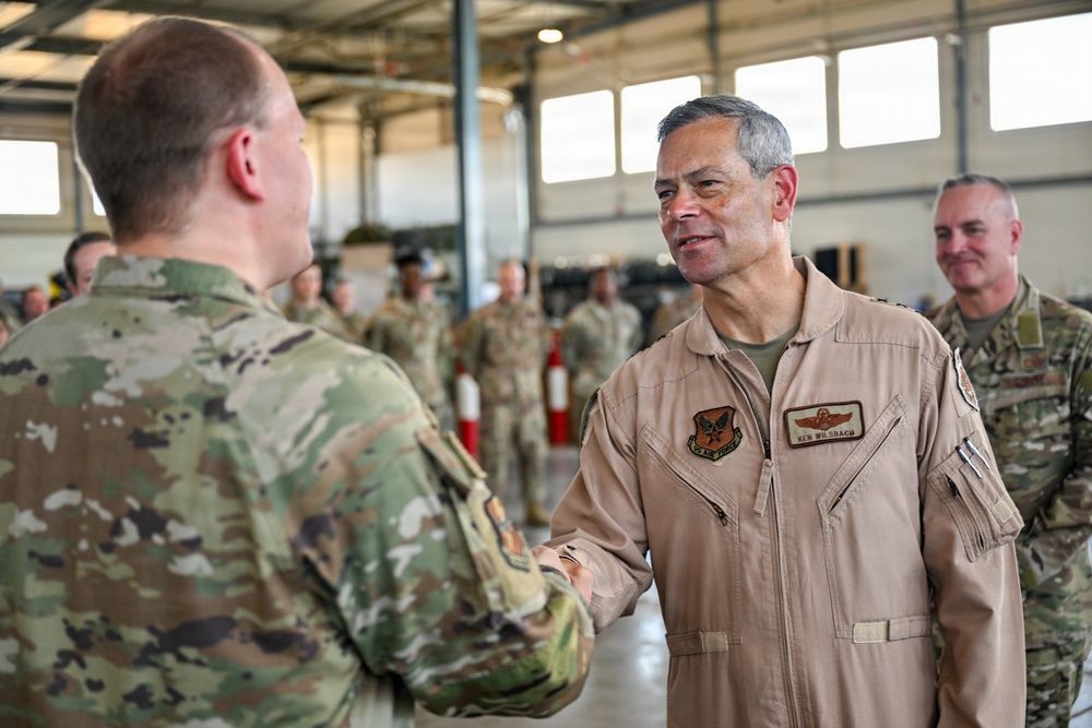 A military officer wearing a tan/khaki flight suit with a name tag reading 'KEN WILSBACH' is shaking hands with another service member in OCP (Operational Camouflage Pattern) uniform inside what appears to be a military hangar. The officer in the flight suit displays pilot wings and a unit patch on his chest. Multiple other uniformed personnel are visible in the background.