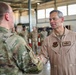 A military officer wearing a tan/khaki flight suit with a name tag reading 'KEN WILSBACH' is shaking hands with another service member in OCP (Operational Camouflage Pattern) uniform inside what appears to be a military hangar. The officer in the flight suit displays pilot wings and a unit patch on his chest. Multiple other uniformed personnel are visible in the background.