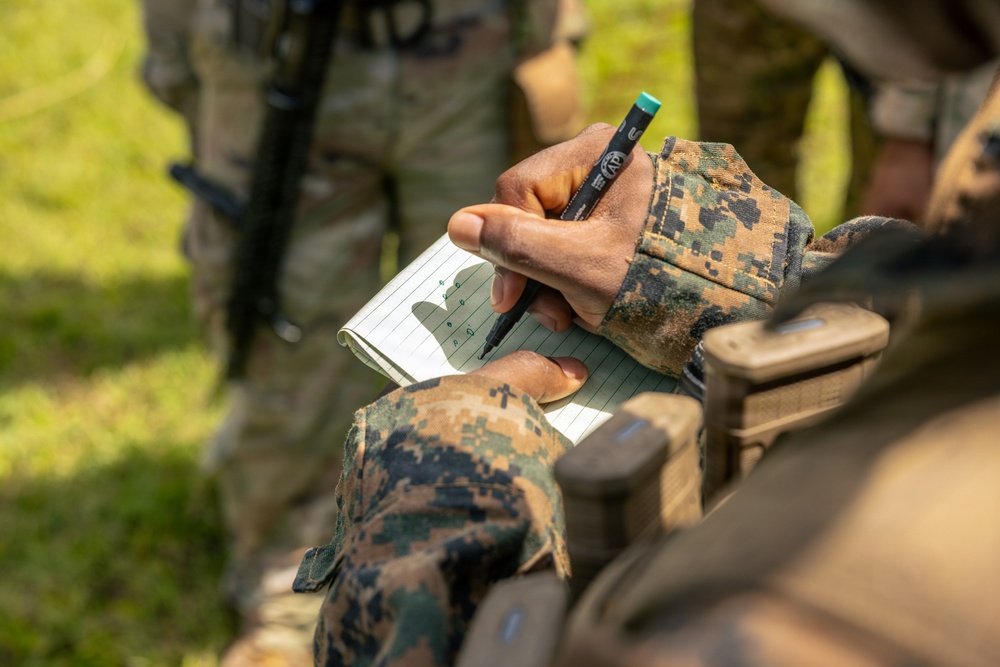A military personnel wearing MARPAT (Marine pattern) digital camouflage uniform is writing notes in a small lined notepad using a black marker with a green cap. The close-up shot focuses on the hands and notepad, with a second uniformed individual visible and blurred in the background. The setting appears to be an outdoor field or training environment.