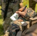 A military personnel wearing MARPAT (Marine pattern) digital camouflage uniform is writing notes in a small lined notepad using a black marker with a green cap. The close-up shot focuses on the hands and notepad, with a second uniformed individual visible and blurred in the background. The setting appears to be an outdoor field or training environment.