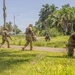 The image shows a group of military personnel conducting what appears to be a tactical training exercise in a tropical environment. Soldiers in camouflage uniforms are moving in formation across a grassy area surrounded by palm trees, with at least one soldier in a kneeling firing position providing cover. The setting suggests a warm, humid climate consistent with a Caribbean or Central American location.