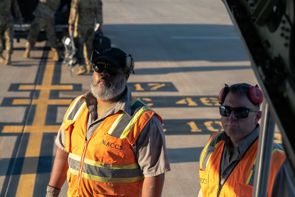 Two ground crew workers wearing orange high-visibility safety vests labeled 'MACCS' are standing on an airfield tarmac. Both men are equipped with hearing protection and sunglasses, suggesting active aircraft operations nearby. Military personnel in camouflage uniforms are visible in the background.