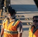 Two ground crew workers wearing orange high-visibility safety vests labeled 'MACCS' are standing on an airfield tarmac. Both men are equipped with hearing protection and sunglasses, suggesting active aircraft operations nearby. Military personnel in camouflage uniforms are visible in the background.