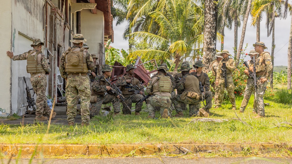 A group of approximately 10-12 armed military personnel in mixed camouflage uniforms are gathered near a weathered building in a tropical environment. Some soldiers are kneeling in a huddle while others stand at the perimeter, suggesting a tactical briefing or coordination exercise. The setting, vegetation, and mixed unit composition indicate a joint military training exercise in a tropical region.