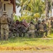 A group of approximately 10-12 armed military personnel in mixed camouflage uniforms are gathered near a weathered building in a tropical environment. Some soldiers are kneeling in a huddle while others stand at the perimeter, suggesting a tactical briefing or coordination exercise. The setting, vegetation, and mixed unit composition indicate a joint military training exercise in a tropical region.
