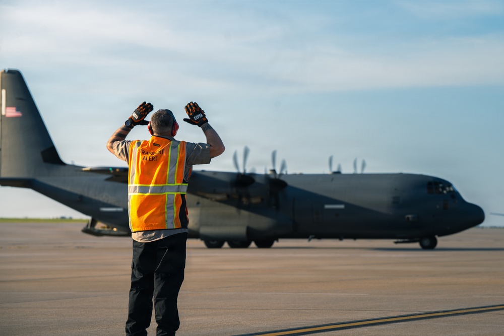 A ground crew member wearing an orange 'Transient Alert' safety vest is marshaling a large military turboprop aircraft, identified as a C-130 Hercules, on a tarmac. The marshalller has both arms raised with gloved hands visible, signaling the aircraft during taxi operations. The image is taken during golden hour lighting, casting warm tones across the scene.