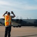 A ground crew member wearing an orange 'Transient Alert' safety vest is marshaling a large military turboprop aircraft, identified as a C-130 Hercules, on a tarmac. The marshalller has both arms raised with gloved hands visible, signaling the aircraft during taxi operations. The image is taken during golden hour lighting, casting warm tones across the scene.