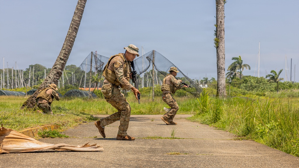Three military personnel in multicam/OCP camouflage uniforms are conducting what appears to be a tactical training exercise on a paved path in a tropical environment. The soldiers are moving in a tactical formation, with two advancing along the path while a third takes a low covered position near a palm tree. The setting includes palm trees, netting structures, and lush tropical vegetation consistent with a Pacific or Caribbean training location.