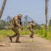 Three military personnel in multicam/OCP camouflage uniforms are conducting what appears to be a tactical training exercise on a paved path in a tropical environment. The soldiers are moving in a tactical formation, with two advancing along the path while a third takes a low covered position near a palm tree. The setting includes palm trees, netting structures, and lush tropical vegetation consistent with a Pacific or Caribbean training location.