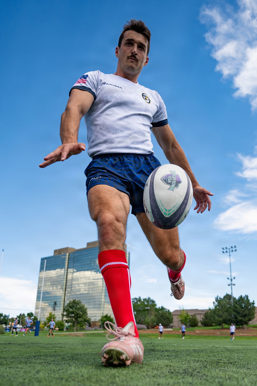 A male rugby player is photographed from a dramatic low-angle ground-level perspective as he prepares to kick a rugby ball. He is wearing a white Akuma jersey with a US flag patch, blue shorts, red socks, and pink cleats. The shot is taken on a green turf field with a modern glass building and blue sky visible in the background.