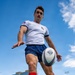 A male rugby player is photographed from a dramatic low-angle ground-level perspective as he prepares to kick a rugby ball. He is wearing a white Akuma jersey with a US flag patch, blue shorts, red socks, and pink cleats. The shot is taken on a green turf field with a modern glass building and blue sky visible in the background.