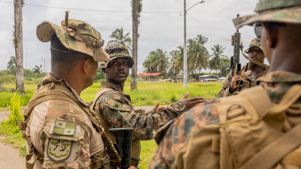 The image shows a group of military personnel in camouflage uniforms engaged in what appears to be a briefing or discussion in an outdoor tropical environment. At least two different military forces are represented, identifiable by distinct uniform patterns and flag patches. The setting includes palm trees, green vegetation, and what appears to be a road or pathway.