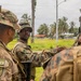 The image shows a group of military personnel in camouflage uniforms engaged in what appears to be a briefing or discussion in an outdoor tropical environment. At least two different military forces are represented, identifiable by distinct uniform patterns and flag patches. The setting includes palm trees, green vegetation, and what appears to be a road or pathway.
