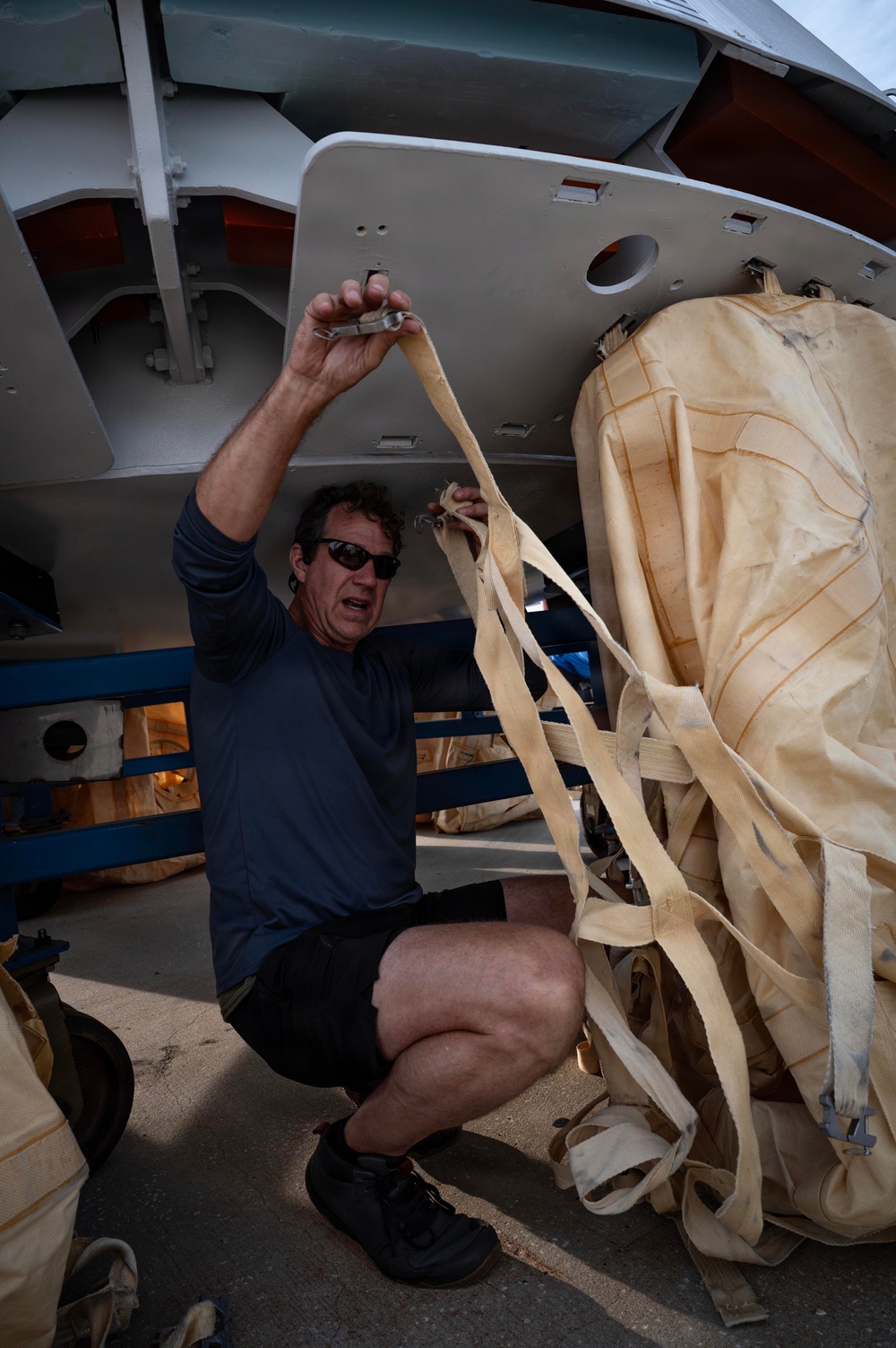A middle-aged man wearing sunglasses, a blue long-sleeve shirt, and black shorts is crouched beneath what appears to be an aircraft fuselage. He is handling straps or webbing connected to a large bundled cargo package wrapped in tan/cream-colored fabric. The setting appears to be an airfield or hangar area with concrete flooring.