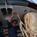 A middle-aged man wearing sunglasses, a blue long-sleeve shirt, and black shorts is crouched beneath what appears to be an aircraft fuselage. He is handling straps or webbing connected to a large bundled cargo package wrapped in tan/cream-colored fabric. The setting appears to be an airfield or hangar area with concrete flooring.