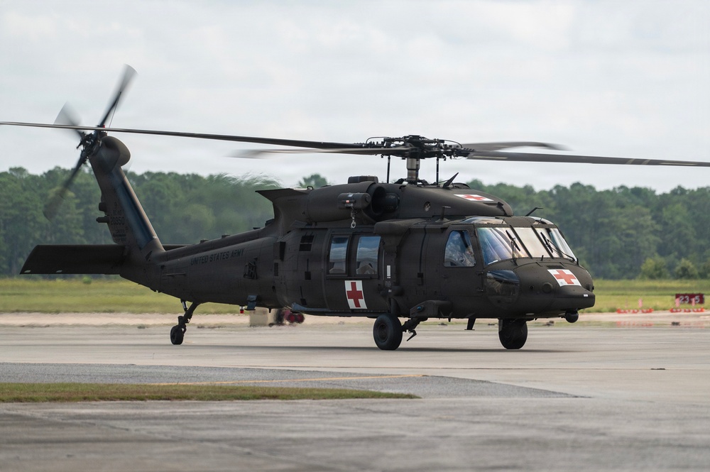 A United States Army UH-60 Black Hawk helicopter configured as a medevac (medical evacuation) aircraft is shown on a tarmac with rotors spinning. The helicopter is painted in olive drab and displays prominent red cross markings on the fuselage and top rotor housing, indicating its medical role. The aircraft appears to be preparing for takeoff or has just landed at a military airfield.