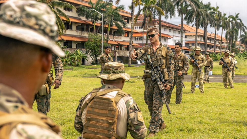 A group of military personnel in mixed camouflage uniforms are gathered on a grassy field near multi-story buildings flanked by palm trees, suggesting a tropical location. A central figure in MARPAT (Marine pattern) camouflage with sunglasses and a rifle appears to be instructing or briefing the group. The scene is consistent with a joint military training exercise involving personnel from multiple nations.