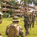 A group of military personnel in mixed camouflage uniforms are gathered on a grassy field near multi-story buildings flanked by palm trees, suggesting a tropical location. A central figure in MARPAT (Marine pattern) camouflage with sunglasses and a rifle appears to be instructing or briefing the group. The scene is consistent with a joint military training exercise involving personnel from multiple nations.