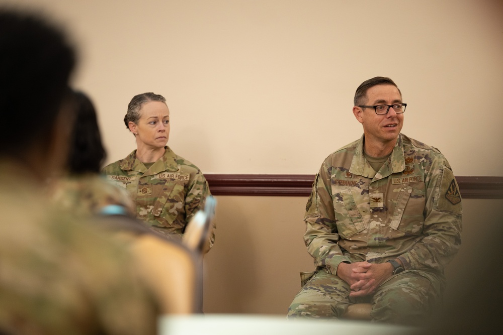 Two U.S. Air Force personnel in OCP (Operational Camouflage Pattern) uniforms are seated and appear to be participating in a meeting or briefing. The individual on the right, wearing glasses, appears to be speaking, while the individual on the left listens attentively. A third partially visible person is seen in the foreground, suggesting an audience or group setting.