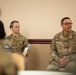 Two U.S. Air Force personnel in OCP (Operational Camouflage Pattern) uniforms are seated and appear to be participating in a meeting or briefing. The individual on the right, wearing glasses, appears to be speaking, while the individual on the left listens attentively. A third partially visible person is seen in the foreground, suggesting an audience or group setting.