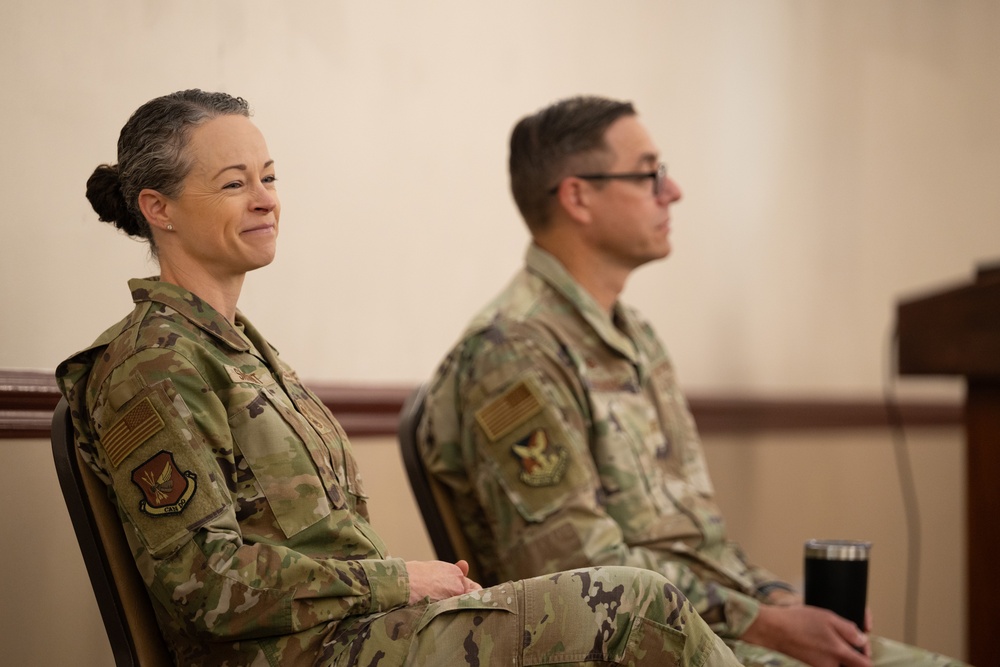 Two U.S. Air Force personnel in Operational Camouflage Pattern (OCP) uniforms are seated side by side, appearing to attend a formal event or briefing. The female service member on the left is smiling, while the male service member on the right is attentively facing forward. Both wear unit patches and U.S. flag patches on their right sleeves.