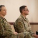 Two U.S. Air Force personnel in Operational Camouflage Pattern (OCP) uniforms are seated side by side, appearing to attend a formal event or briefing. The female service member on the left is smiling, while the male service member on the right is attentively facing forward. Both wear unit patches and U.S. flag patches on their right sleeves.