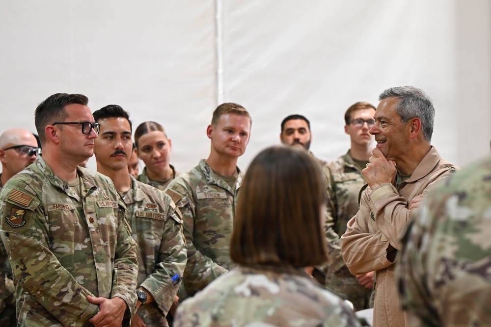 A group of U.S. Air Force personnel in OCP (Operational Camouflage Pattern) uniforms are gathered inside what appears to be a tent or temporary structure. A senior military officer in a tan/desert flight suit is addressing or conversing with the assembled airmen. The setting suggests a briefing, visit, or informal meeting at a forward operating location.