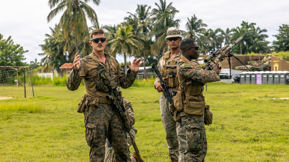 Three military personnel in camouflage uniforms are gathered on a grassy field in what appears to be a tropical location, identifiable by the palm trees in the background. One soldier on the left appears to be briefing or instructing the other two, gesturing with both hands. The soldiers are fully equipped with tactical gear, body armor, and rifles.