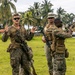 Three military personnel in camouflage uniforms are gathered on a grassy field in what appears to be a tropical location, identifiable by the palm trees in the background. One soldier on the left appears to be briefing or instructing the other two, gesturing with both hands. The soldiers are fully equipped with tactical gear, body armor, and rifles.