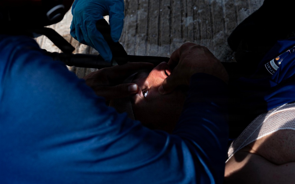 A person is lying on what appears to be a stretcher or backboard receiving medical attention. A medical responder wearing blue latex gloves is securing straps or providing care to the patient's head and neck area. The scene suggests an emergency medical response situation.