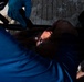A person is lying on what appears to be a stretcher or backboard receiving medical attention. A medical responder wearing blue latex gloves is securing straps or providing care to the patient's head and neck area. The scene suggests an emergency medical response situation.
