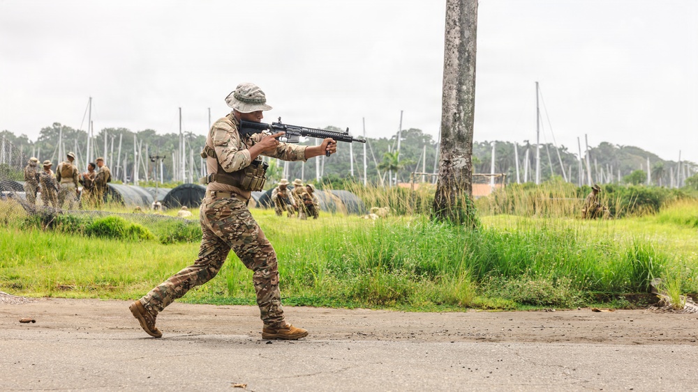 A soldier in camouflage uniform and boonie hat is moving in a tactical stance while aiming a rifle on a paved surface. In the background, multiple other military personnel are visible engaged in what appears to be a training exercise in an open field environment. The setting appears to be a military training area with lush green vegetation and overcast skies.