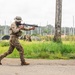 A soldier in camouflage uniform and boonie hat is moving in a tactical stance while aiming a rifle on a paved surface. In the background, multiple other military personnel are visible engaged in what appears to be a training exercise in an open field environment. The setting appears to be a military training area with lush green vegetation and overcast skies.