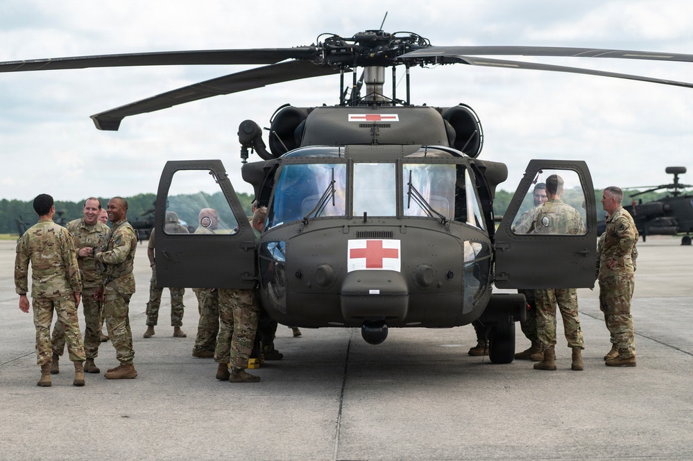 A group of military personnel in camouflage uniforms are gathered around a U.S. Army UH-60 Black Hawk helicopter configured as a medevac (medical evacuation) aircraft, identifiable by the prominent Red Cross markings on both the fuselage and rotor housing. The scene takes place on a military airfield tarmac, with additional helicopters visible in the background. Personnel appear to be engaged in conversation and inspection of the aircraft.