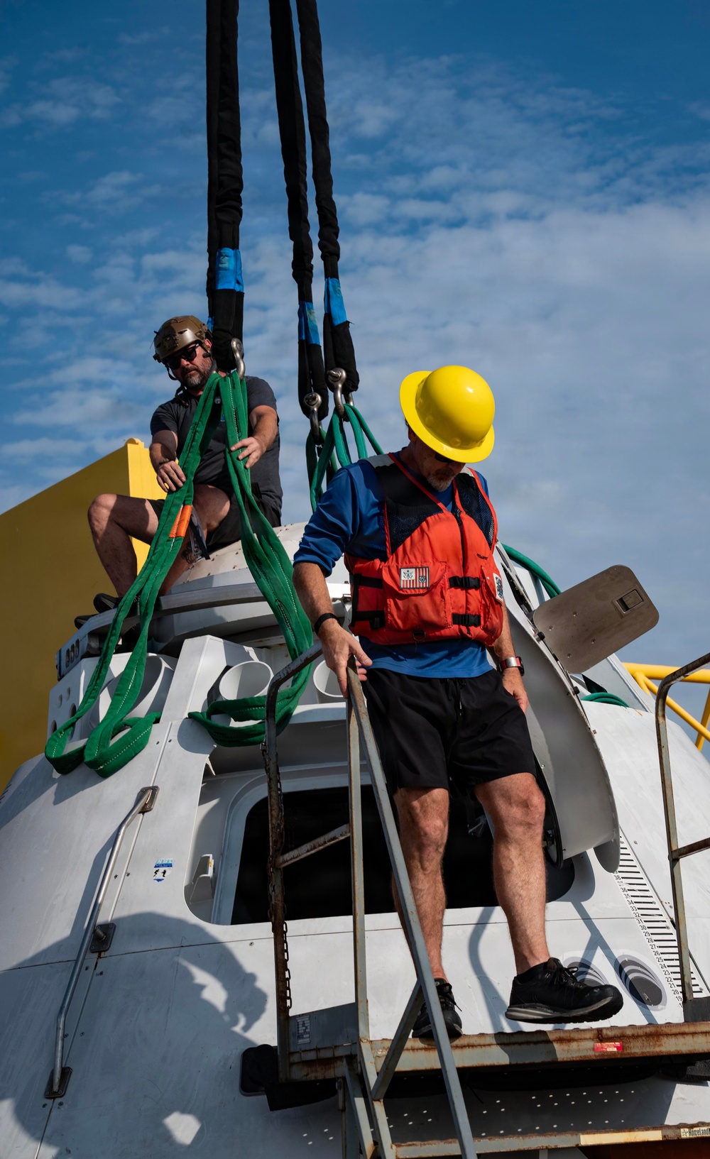 Two workers are handling a large white capsule-shaped object, likely a spacecraft or recovery capsule, using green lifting straps and black crane rigging. One worker wearing a yellow hard hat and orange life vest is descending a ladder on the side of the capsule, while a second worker in a tan helmet sits atop the capsule managing the green straps. The scene appears to be a maritime recovery or rigging operation conducted on a vessel deck.