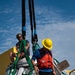 Two workers are handling a large white capsule-shaped object, likely a spacecraft or recovery capsule, using green lifting straps and black crane rigging. One worker wearing a yellow hard hat and orange life vest is descending a ladder on the side of the capsule, while a second worker in a tan helmet sits atop the capsule managing the green straps. The scene appears to be a maritime recovery or rigging operation conducted on a vessel deck.