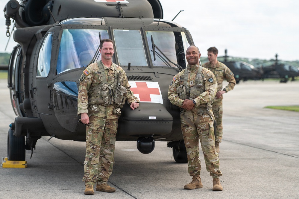 Two U.S. Army soldiers in MultiCam (OCP) camouflage uniforms pose in front of a UH-60 Black Hawk medevac helicopter marked with a red cross, indicating medical evacuation (MEDEVAC) designation. A third soldier stands in the background near additional military helicopters on what appears to be a military airfield or flight line. The image appears to be an official or semi-official military photograph.