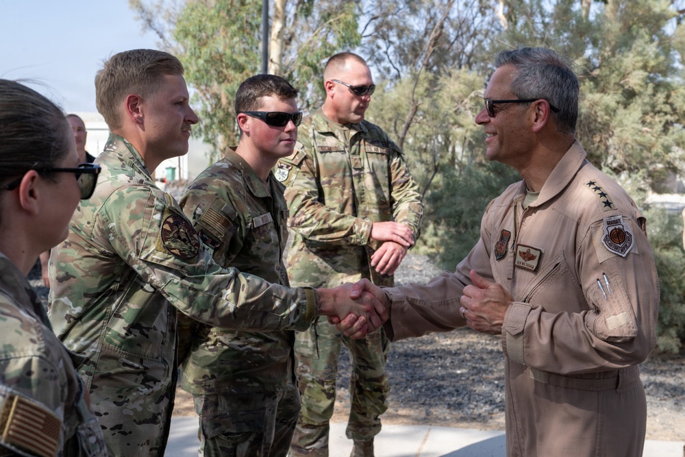 A senior military officer in a tan/khaki flight suit is greeting and shaking hands with several service members dressed in OCP (Operational Camouflage Pattern) uniforms. The setting appears to be an outdoor military installation, likely in an arid region based on the vegetation and environment. The officer on the right displays what appear to be general officer rank insignia (four stars visible on collar area).