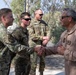 A senior military officer in a tan/khaki flight suit is greeting and shaking hands with several service members dressed in OCP (Operational Camouflage Pattern) uniforms. The setting appears to be an outdoor military installation, likely in an arid region based on the vegetation and environment. The officer on the right displays what appear to be general officer rank insignia (four stars visible on collar area).