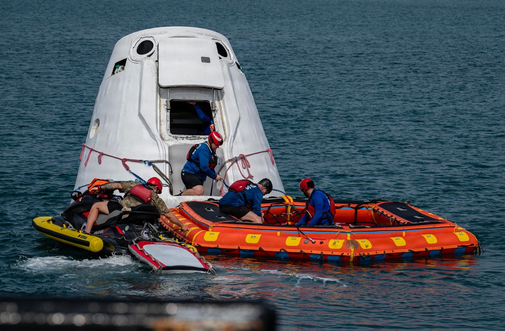Recovery personnel are working alongside a SpaceX Dragon capsule floating in open ocean water following a splashdown. A team of at least five workers in safety gear are operating from an orange inflatable rescue raft and a yellow personal watercraft (jet ski) to secure and access the capsule. The capsule's hatch appears to be open, suggesting crew extraction or post-landing procedures are underway.
