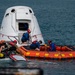 Recovery personnel are working alongside a SpaceX Dragon capsule floating in open ocean water following a splashdown. A team of at least five workers in safety gear are operating from an orange inflatable rescue raft and a yellow personal watercraft (jet ski) to secure and access the capsule. The capsule's hatch appears to be open, suggesting crew extraction or post-landing procedures are underway.