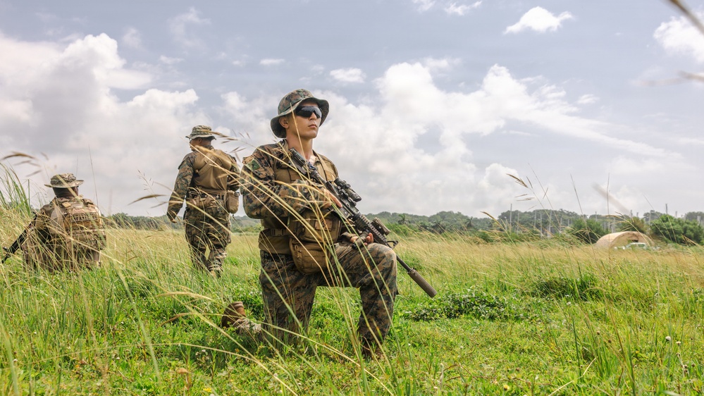 Three military personnel in MARPAT woodland/desert camouflage uniforms are moving through an open grassy field. The foreground soldier is kneeling and holding a scoped rifle while scanning the surroundings, with two others walking behind. The setting appears to be a training exercise based on the open terrain and visible tent structures in the background.