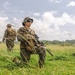 Three military personnel in MARPAT woodland/desert camouflage uniforms are moving through an open grassy field. The foreground soldier is kneeling and holding a scoped rifle while scanning the surroundings, with two others walking behind. The setting appears to be a training exercise based on the open terrain and visible tent structures in the background.
