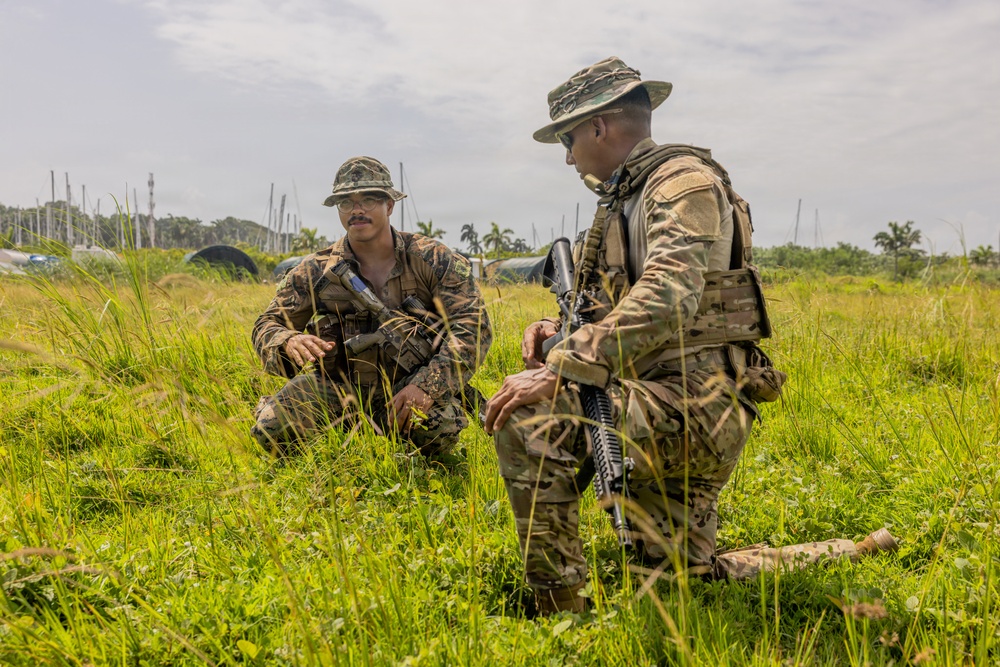 Two armed military personnel in camouflage uniforms are kneeling in a grassy field, appearing to be in conversation or conducting a briefing. Both individuals are equipped with tactical gear, weapons, and boonie hats, suggesting a field training exercise or operational activity. The setting appears to be a tropical or subtropical environment based on the vegetation and palm trees visible in the background.