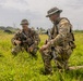 Two armed military personnel in camouflage uniforms are kneeling in a grassy field, appearing to be in conversation or conducting a briefing. Both individuals are equipped with tactical gear, weapons, and boonie hats, suggesting a field training exercise or operational activity. The setting appears to be a tropical or subtropical environment based on the vegetation and palm trees visible in the background.